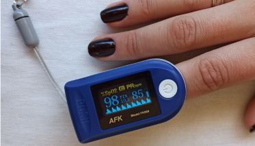 Fotografia em close-up de um oxímetro de pulso digital portátil, de cor azul, fixado no dedo indicador de uma mão com unhas pintadas de esmalte preto. O visor do aparelho está ligado e exibe o valor de saturação de oxigênio (SpO2) em 98% e a frequência cardíaca em 85 batimentos por minuto (PRbpm), junto a uma curva pletismográfica azul. O fundo é de tecido branco.
