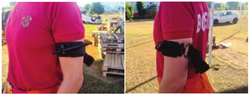 Duas fotografias lado a lado demonstrando a aplicação de um torniquete tático no braço esquerdo de um indivíduo vestindo uma camiseta polo vermelha. Na foto da esquerda, em vista lateral, destaca-se o torniquete preto posicionado no terço superior do braço com a haste de torção (molinete) fixada. Na foto da direita, em vista posterior oblíqua, observa-se o sistema de fivela e o fechamento do mesmo torniquete. O cenário ao fundo é uma área externa com gramado.